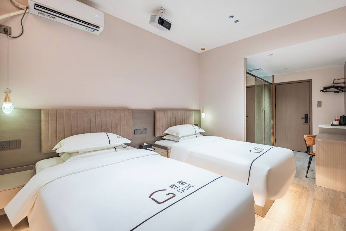 A hotel room with two beds, a desk, a chair, an air conditioning unit, and a projector on the ceiling.