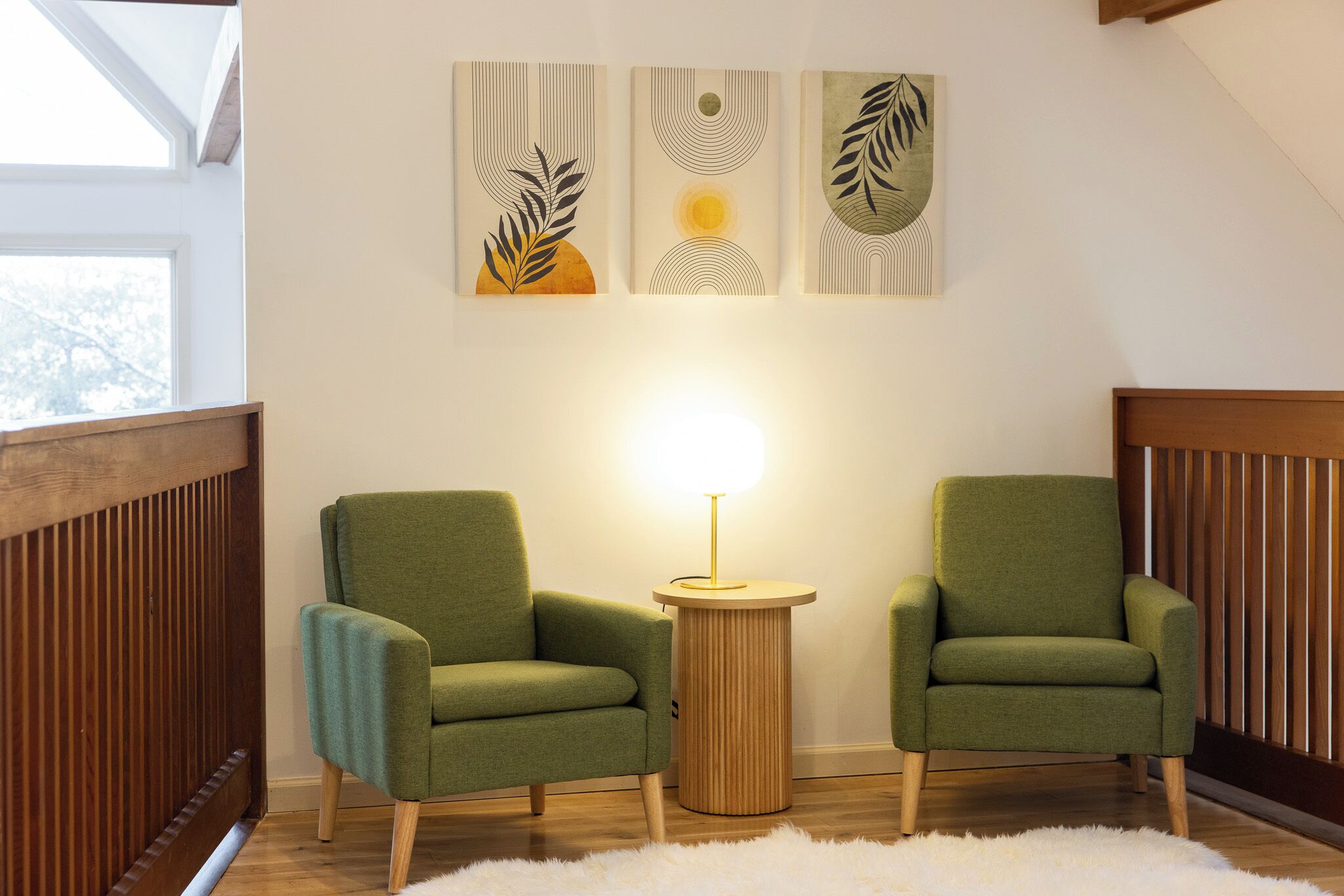 Een kamer met twee groene fauteuils, een rond houten bijzettafeltje met een lamp, en drie abstracte kunstwerken aan de muur.