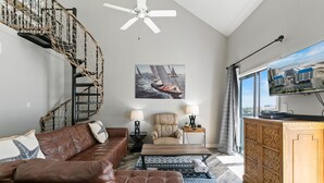 Un salon avec un canapé en cuir marron, un tapis à motifs, une table basse en bois, un fauteuil beige, un téléviseur posé sur un meuble en bois et un escalier en colimaçon.