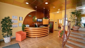 A reception area with a wooden reception desk, potted plants, a staircase, and a visible exit sign. - Aparthotel Pineda (San Michele al Tagliamento)