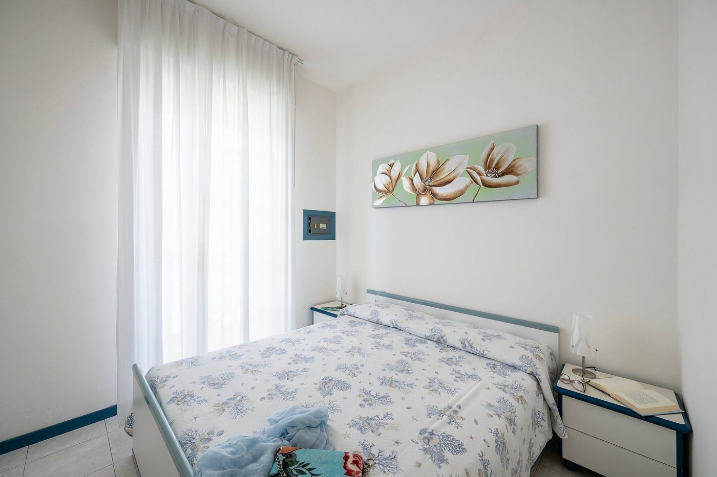 Quarto com cama, mesa de cabeceira, cortinas na janela e uma obra de arte na parede.