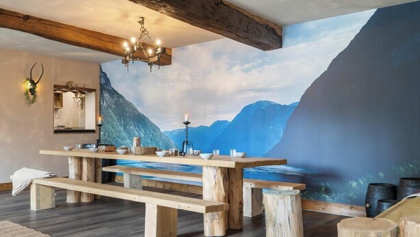 Ein rustikaler Essbereich mit einem Holztisch, Bänken, einer Kristallleuchter und einer Wandmalerei mit Berglandschaft.