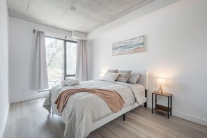 Ein Schlafzimmer mit einem Bett, einem Nachttisch mit Lampe, einem Fenster mit Vorhängen und einem Bild an der Wand.