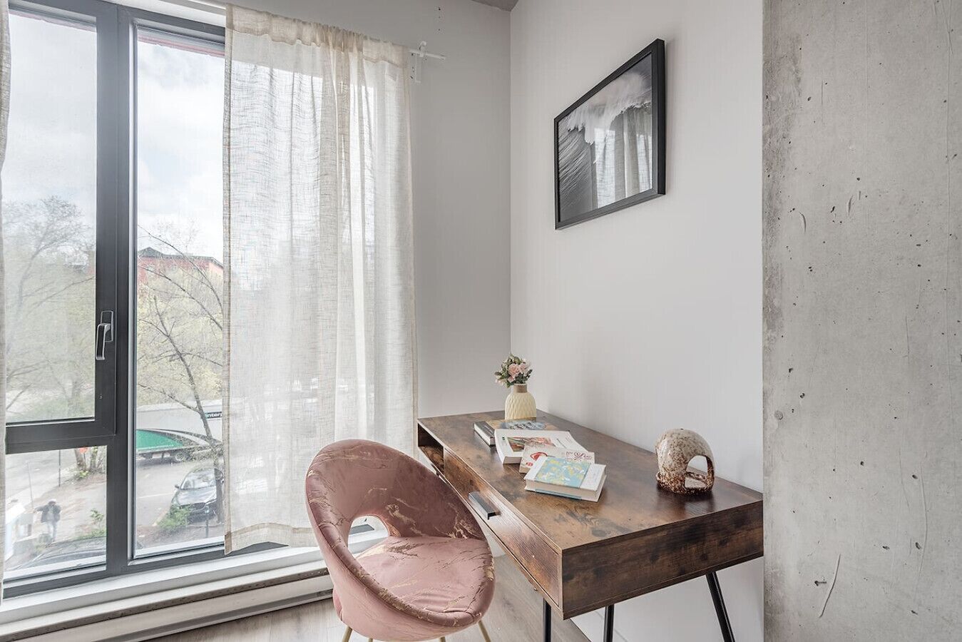 A room with a large window, a wooden desk, a pink chair, and a framed picture on the wall.