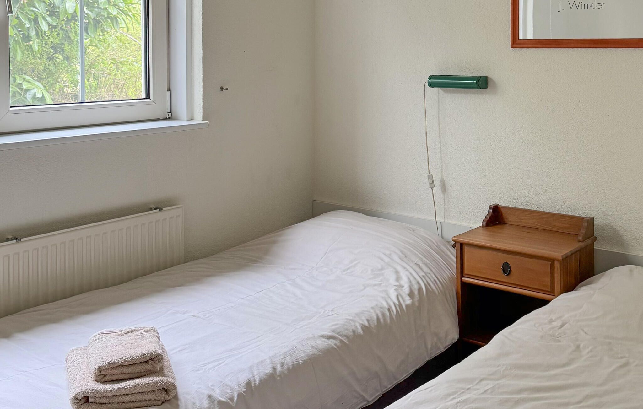 A simple, clean room with a single bed, a nightstand, a radiator, and a window with a view of greenery.