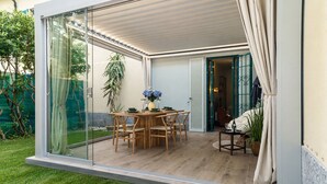 Un espacio moderno para comer al aire libre con pared de vidrio, muebles de madera y una planta en maceta.
