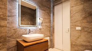 Een badkamer met een marmeren wand, een wit wastafel en een houten opbergmeubel, en een haardroger bevestigd aan de muur.