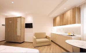 A hotel room with a bed, a sofa, a TV, a kitchenette, and wooden cabinets. - Cozy Apartment 06 in Durr s (Durrës)