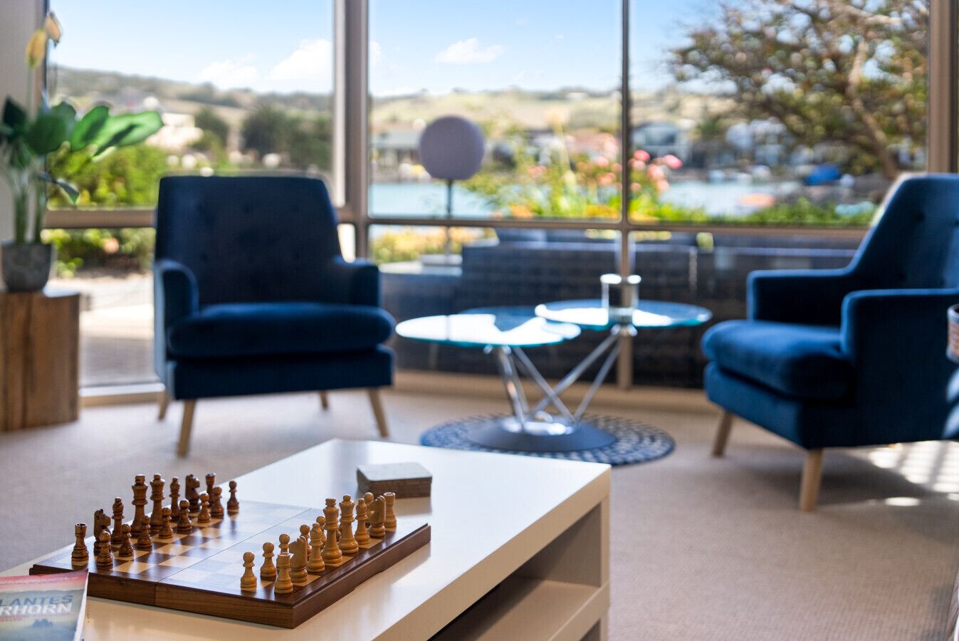 A chessboard on a table, two blue armchairs, a round table, and a view of a lake through large windows.
