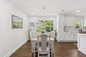 A dining area with a white table and chairs, a chandelier, and a window with a view of greenery. - Pet Friendly Retreat | 3BR w/ Sunroom, Spacious Kitchen + Close to Beach (Saint Simons Island)