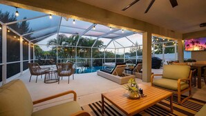 A covered outdoor area with seating, a pool, and a TV screen displaying a sunset. - Infinity Escape - 3BR with Boat Dock and Southern Exposure (Cape Coral)