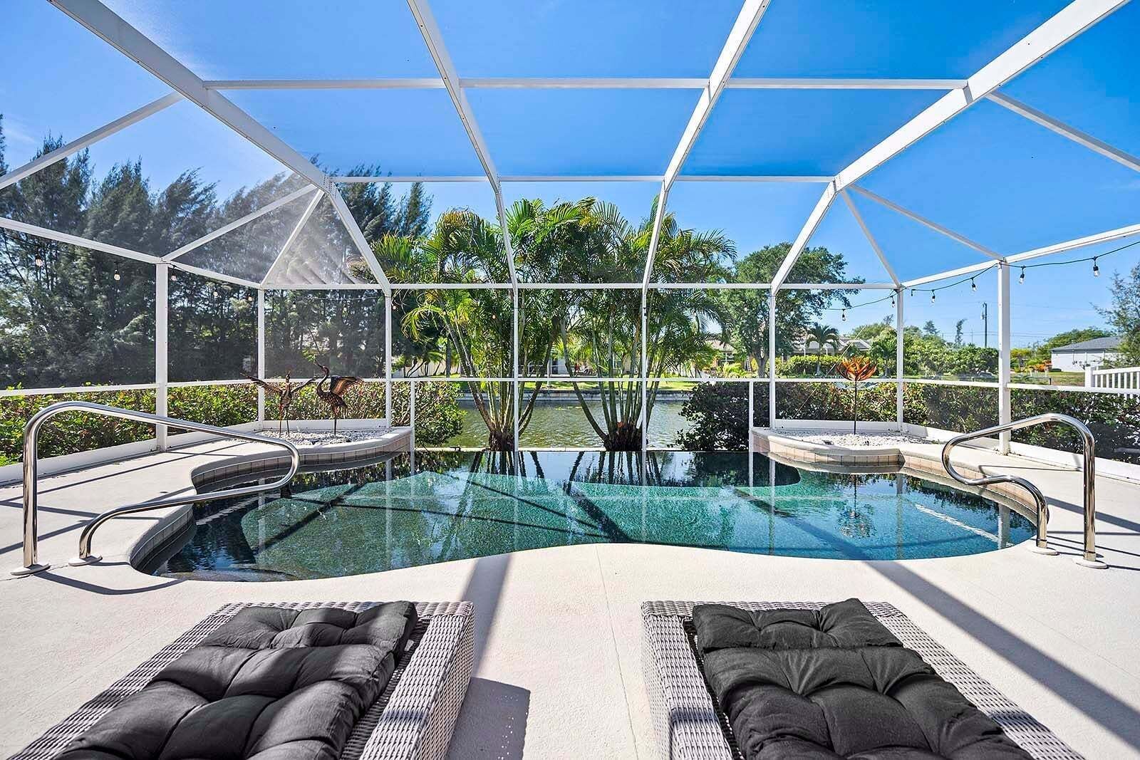 A pool with a glass enclosure, surrounded by lounge chairs and a view of trees and a body of water.