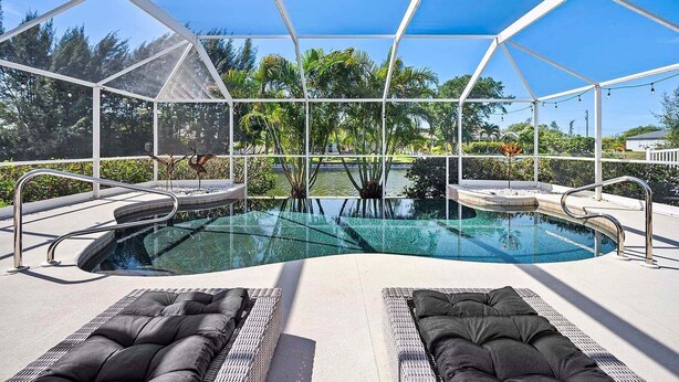 A pool with a glass enclosure, surrounded by lounge chairs and a view of trees and a body of water. - Infinity Escape - 3BR with Boat Dock and Southern Exposure (Cape Coral)
