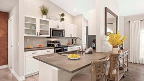 A modern kitchen with white cabinetry, a central island, and a dining area with chairs. - Infinity Escape - 3BR with Boat Dock and Southern Exposure (Cape Coral)