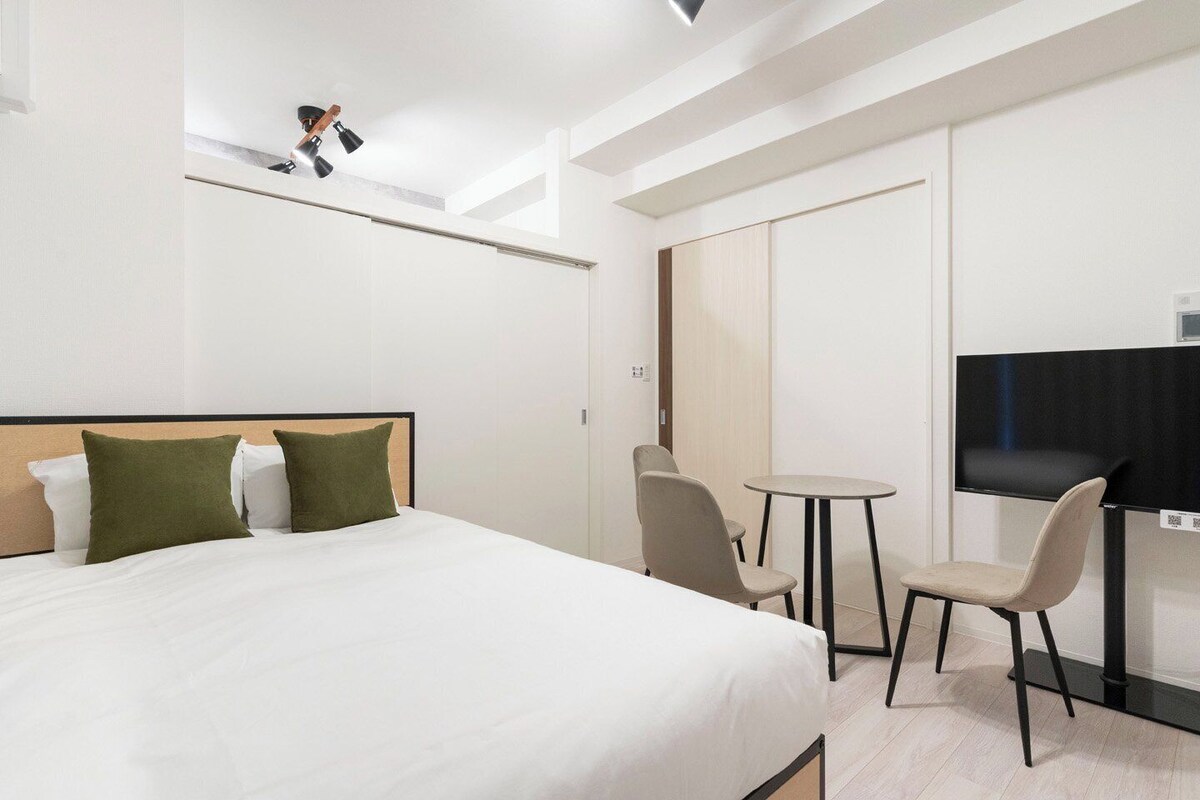 A modern hotel room with a bed, two chairs, a small table, a desk, and a mirror.