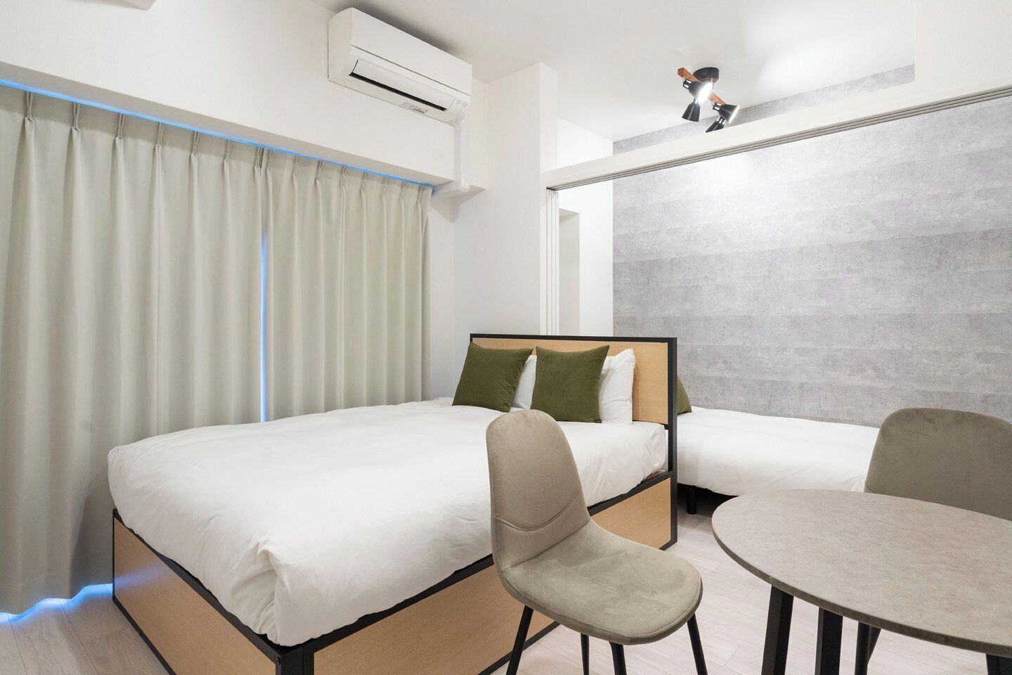 A hotel room with a bed, two chairs, a table, an air conditioning unit, and a wall-mounted lamp.