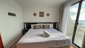 Habitación de hotel con una cama grande, dos mesitas de noche, vistas al mar y una ventana con cortinas.