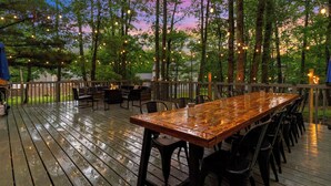 An outdoor deck with a wooden table and chairs, surrounded by trees and string lights. - 5BR Resort-Style - Game Room, Hot Tub, Fire Pit (East Stroudsburg)