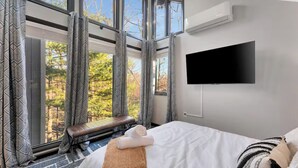 A modern hotel room with large windows, a flat-screen TV, a bed with white linens, and a bench by the window. - 5BR Resort-Style - Game Room, Hot Tub, Fire Pit (East Stroudsburg)