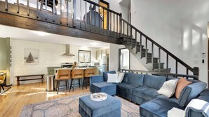A spacious living area with a large sectional sofa, a coffee table, and a patterned rug. A kitchen with a dining area is visible in the background. - 5BR Resort-Style - Game Room, Hot Tub, Fire Pit (East Stroudsburg)
