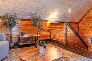 A cozy attic room with wooden walls and a sloped ceiling. There is a table with a plant and puzzle pieces, a couch, and a staircase leading to the upper floor. - Just Remodeled Cabin w/ 4-Game Arcade, Hot Tub & Peloton | 4 Mi to Dollywood! (Pigeon Forge)