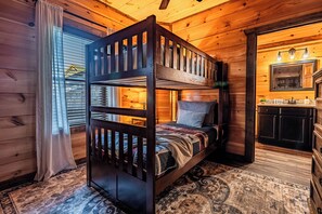 Uma cama beliche com estrutura de madeira, um tapete estampado e vista para uma área de cozinha com espelho e armários.