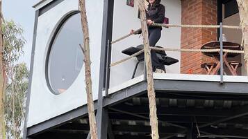 Una casa en un árbol con un ventanal circular grande, una chimenea de ladrillo y una escalera de cuerda.