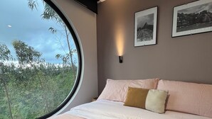 Un dormitorio con un ventanal grande, una cama con almohadas, una lámpara fijada a la pared y dos cuadros enmarcados.