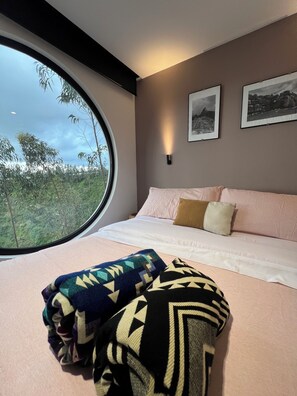 A bedroom with a large window, a bed with pillows, a wall-mounted light, and two framed pictures.