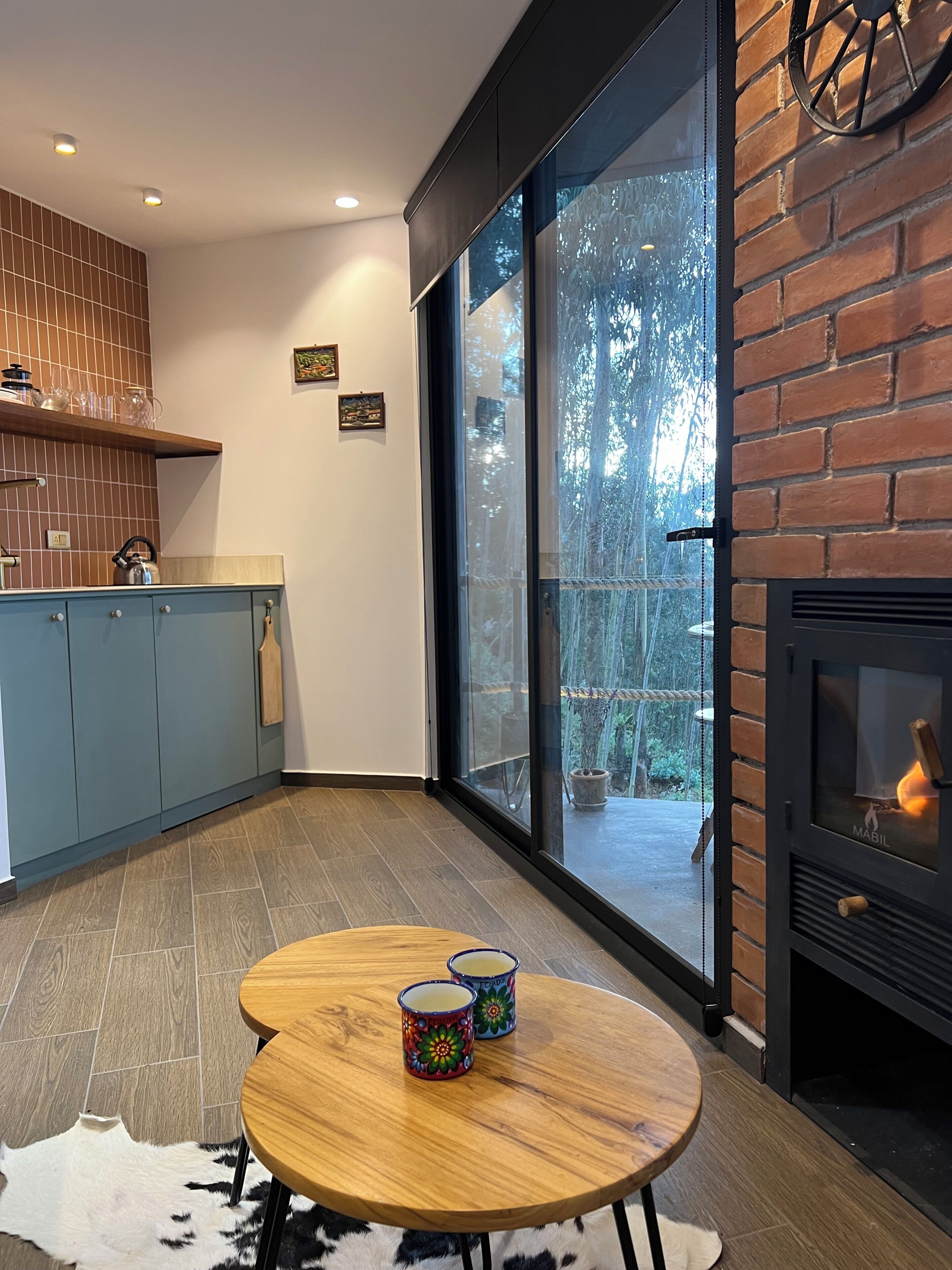Una cocina moderna con un armario azul, una mesa de madera con dos tazas coloridas, una chimenea y un ventanal con vistas a los árboles.