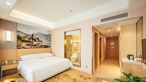 A hotel room with a bed, bedside table, a painting, a mirror, a bathroom, and a kitchenette. - Vienna 3 Good Hotel Huaran Branch (Xiangxi)