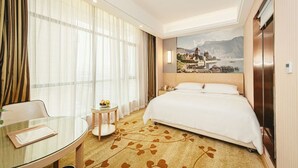 A hotel room with a bed, a glass table, a small round table with fruit, a painting of a landscape, and a view of a body of water. - Vienna 3 Good Hotel Huaran Branch (Xiangxi)