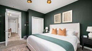 A bedroom with a large bed, a nightstand with a lamp, and a door leading to another room. - Steps to Mass Ave • Sleeps 9 • Free Parking (Indianapolis)