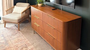 Ein Zimmer mit einem Holzkasten, einem Sessel, einer Topfpflanze, einer Lampe und einem flachbildfernseher, der an der Wand befestigt ist.