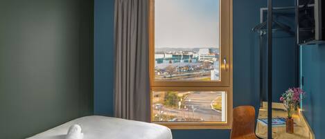 Habitación de hotel con cama, una silla, una mesita con un jarrón y ventana con vista al aeropuerto.