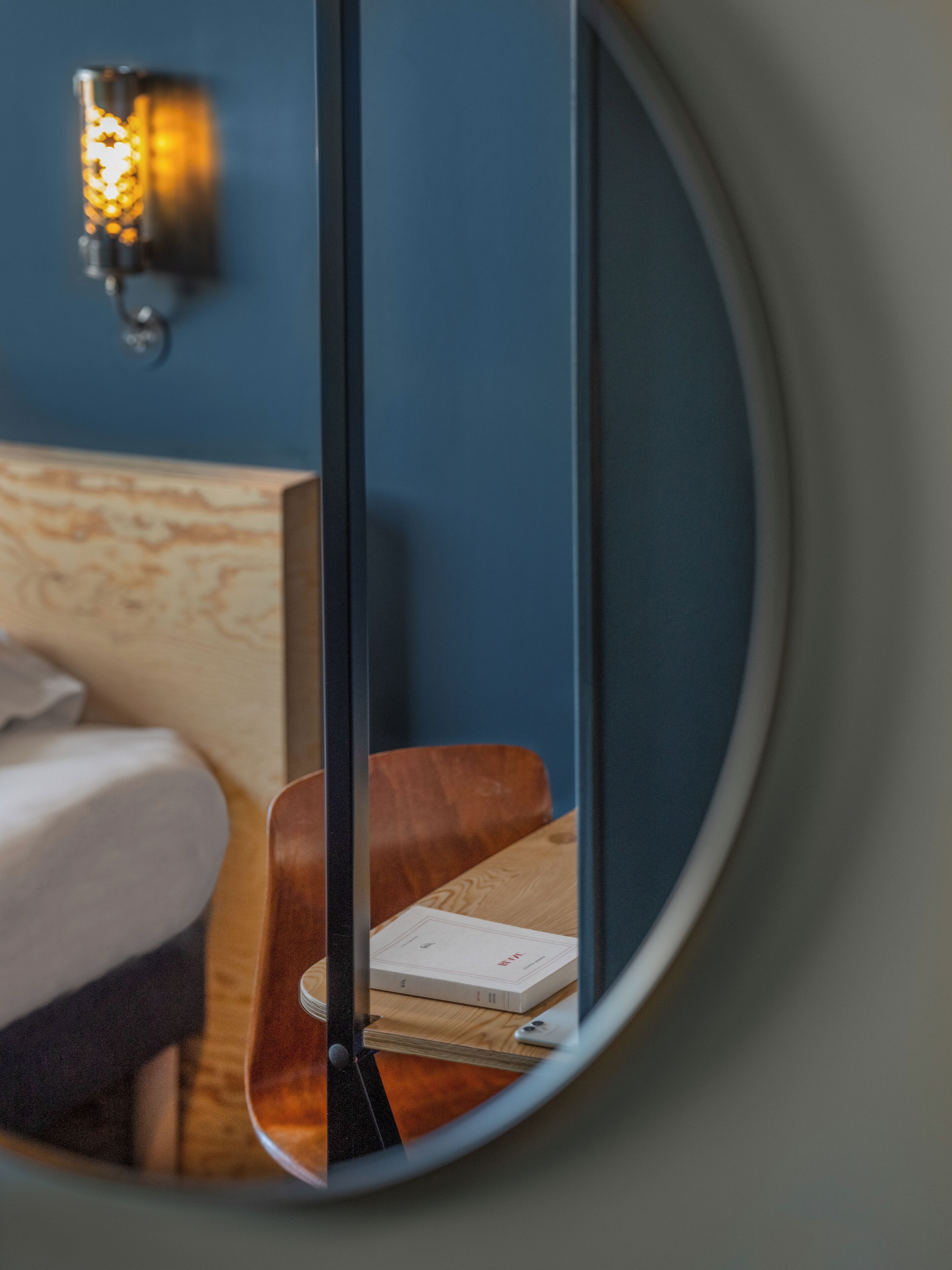 A circular mirror reflecting a room with a bed, a wooden nightstand, a lamp, and a book.