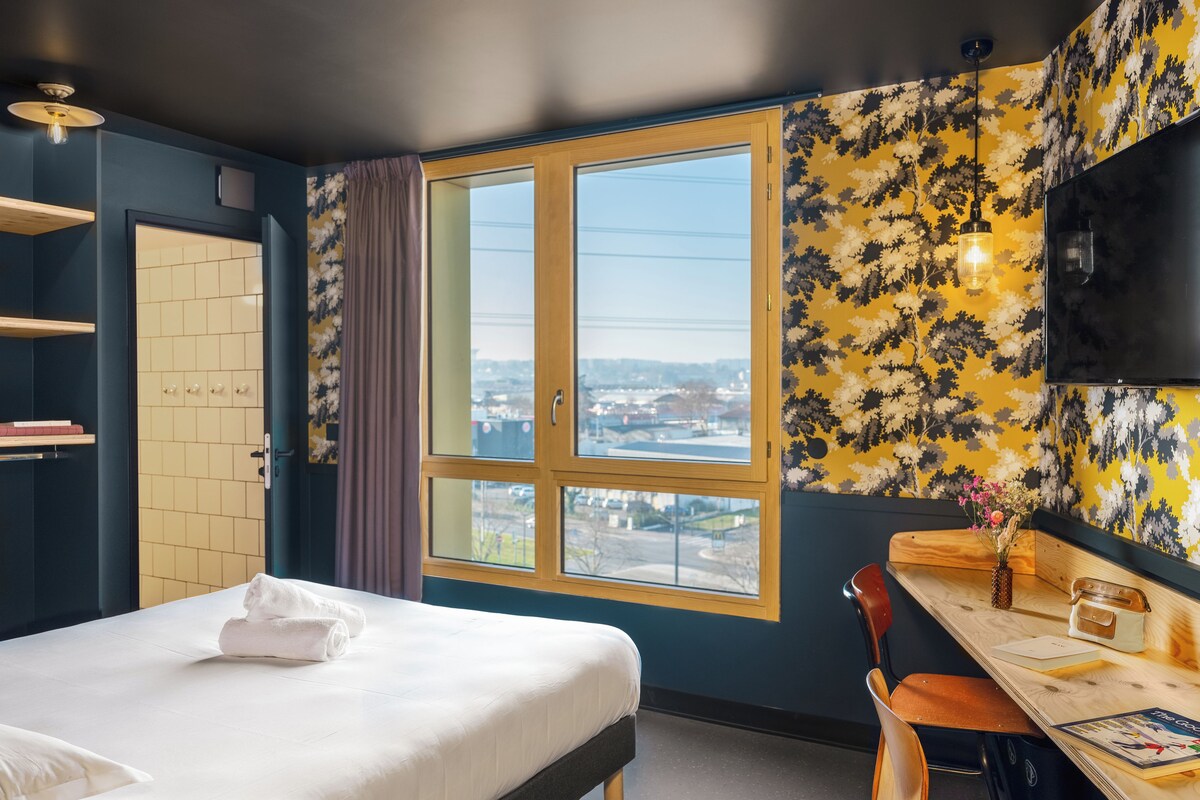A hotel room with a bed, a desk, a chair, a TV, and a window with a city view.