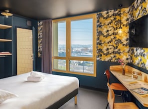 A hotel room with a bed, a desk, a chair, a TV, a window with a city view, and a door with a towel on the bed. - Eklo Hotels Montpellier Centre Gare (Montpellier)