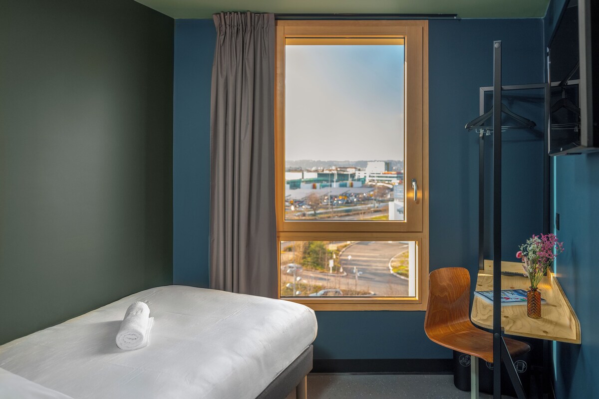 A hotel room with a bed, a chair, a small table with a vase, and a window with a view of an airport.