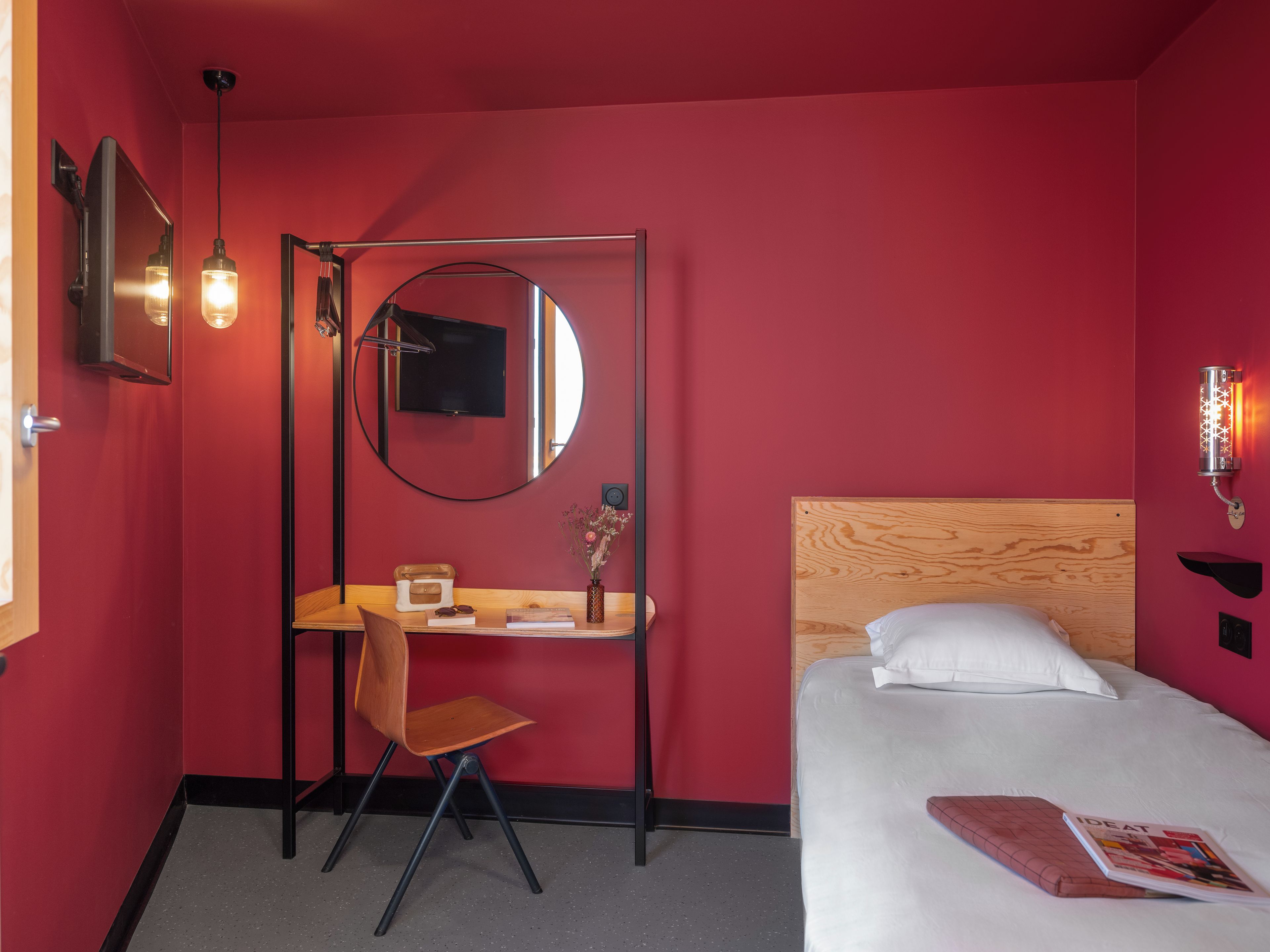 Una habitación de hotel con un escritorio de madera, una silla, una cama con ropa de cama blanca, un espejo redondo, un televisor de pantalla plana y una lámpara de pared.