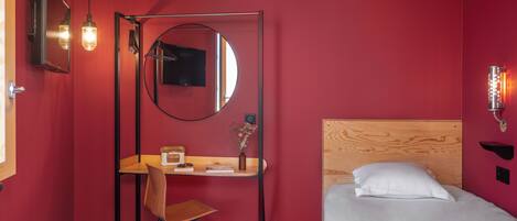 Una habitación de hotel con un escritorio de madera, una silla, una cama con ropa de cama blanca, un espejo redondo, un televisor de pantalla plana y una lámpara de pared.