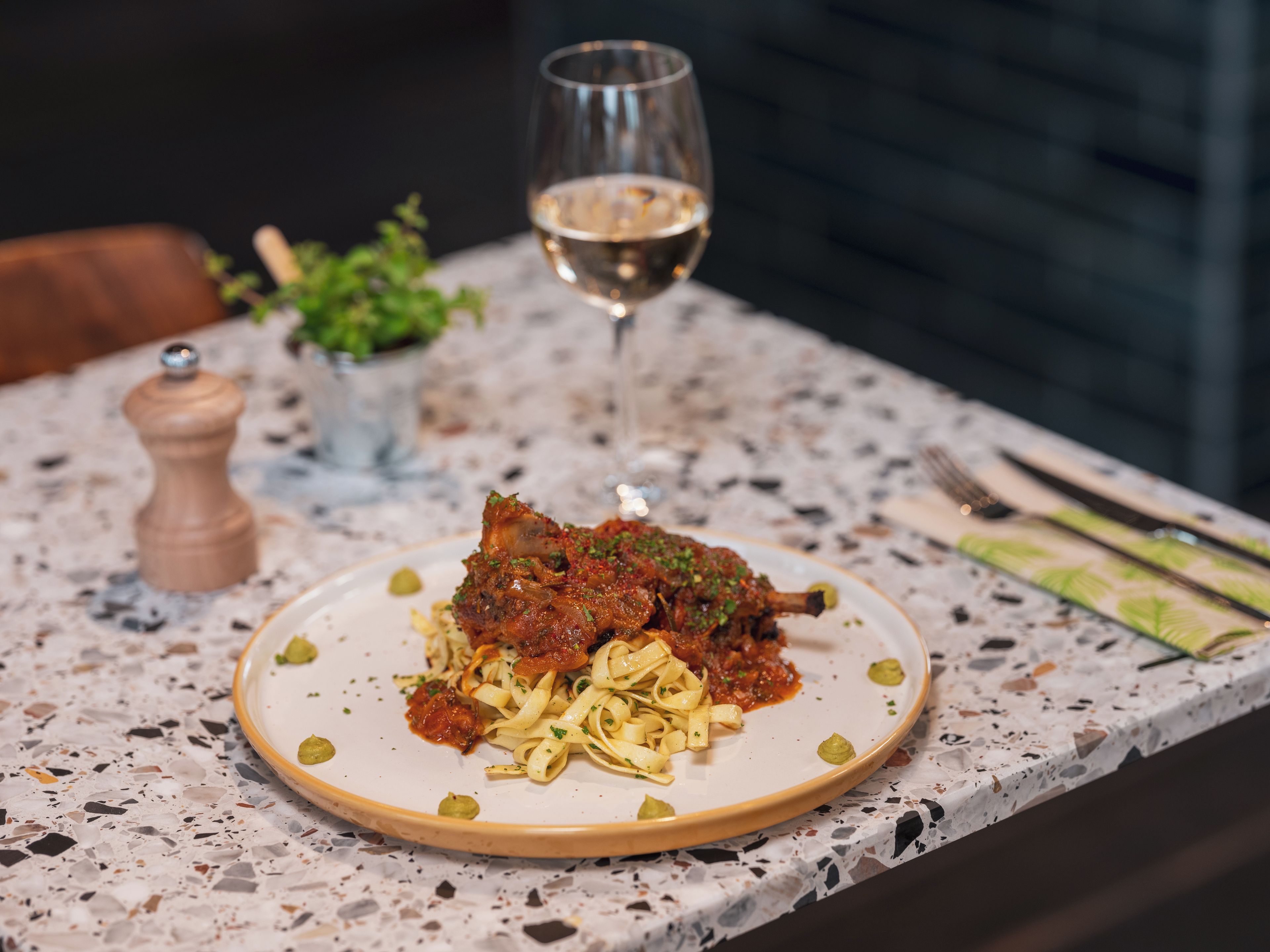 A plate of pasta with a meat topping, a glass of white wine, and a salt and pepper shaker on a speckled countertop.