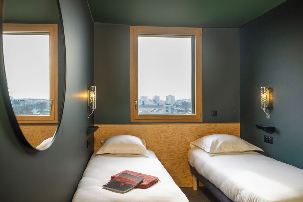 A hotel room with two beds, large windows, and wall-mounted light fixtures.