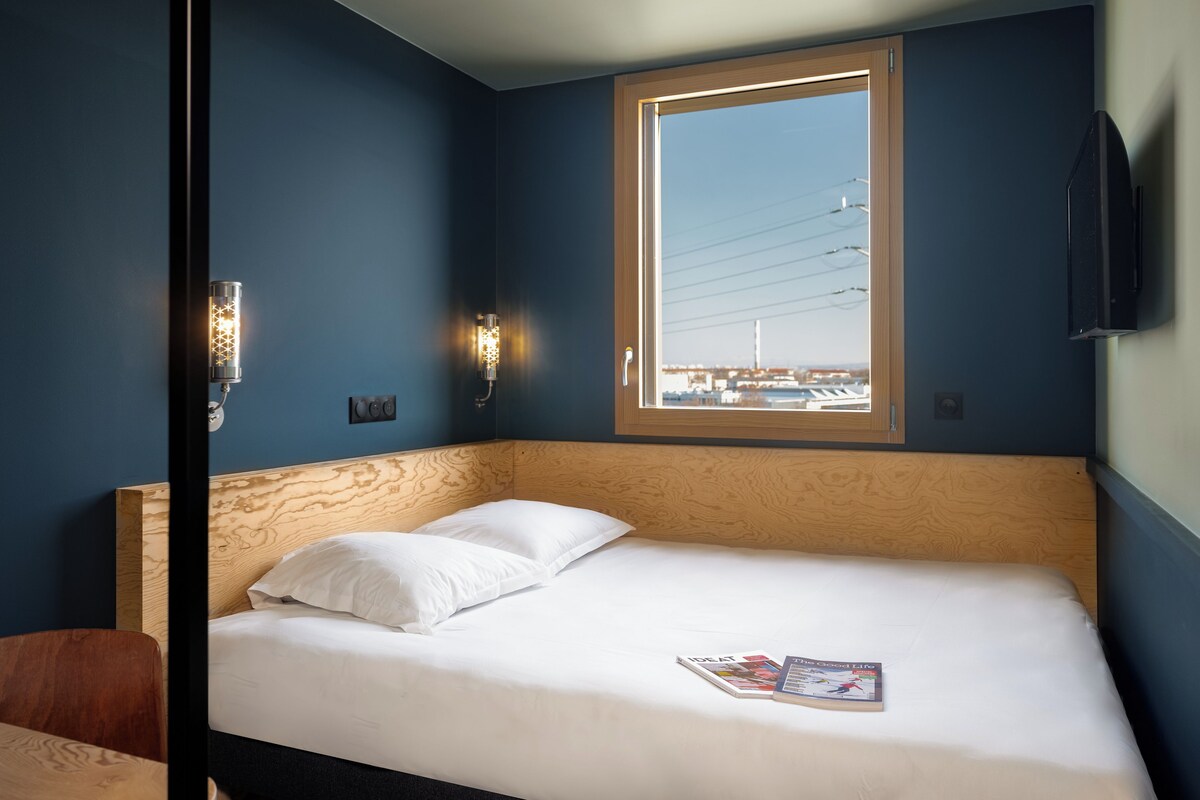 A hotel room with a bed, two pillows, a magazine, a window with a view of buildings, and a wall-mounted TV.