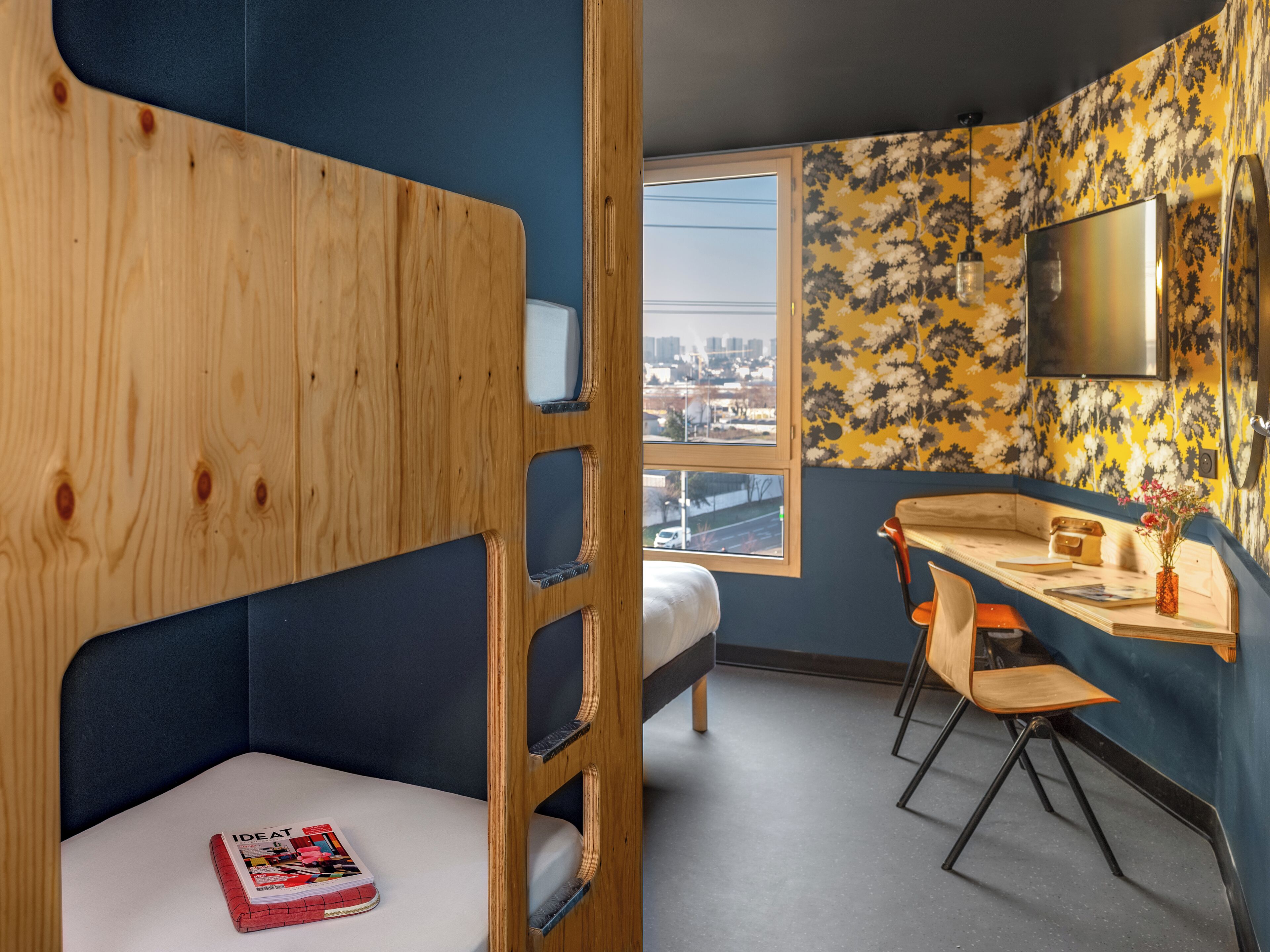 A room with bunk beds, a desk, a chair, and a window with a city view.