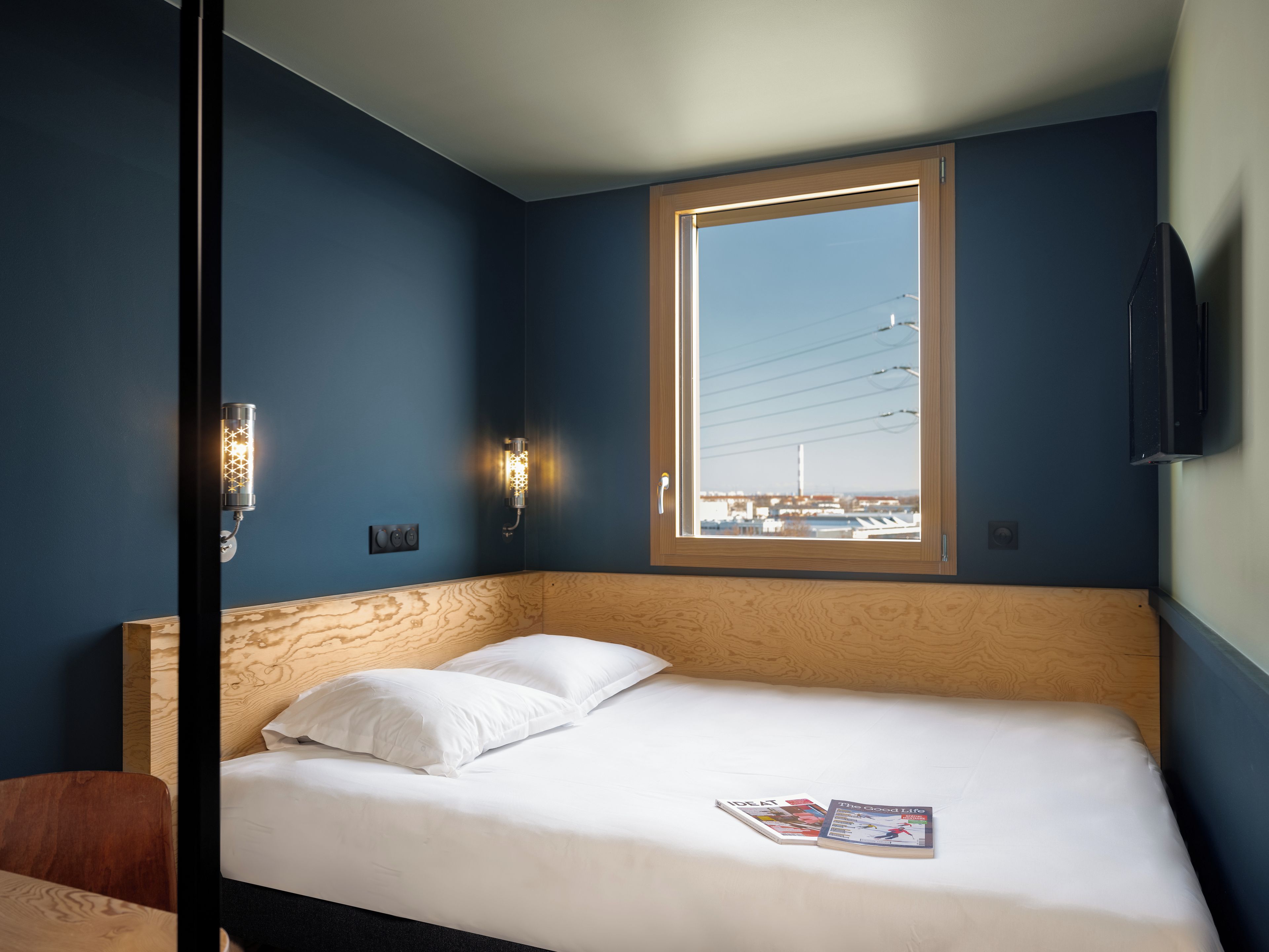 Une chambre d’hôtel avec un lit, deux oreillers, un magazine, une fenêtre donnant sur des immeubles et un téléviseur fixé au mur.