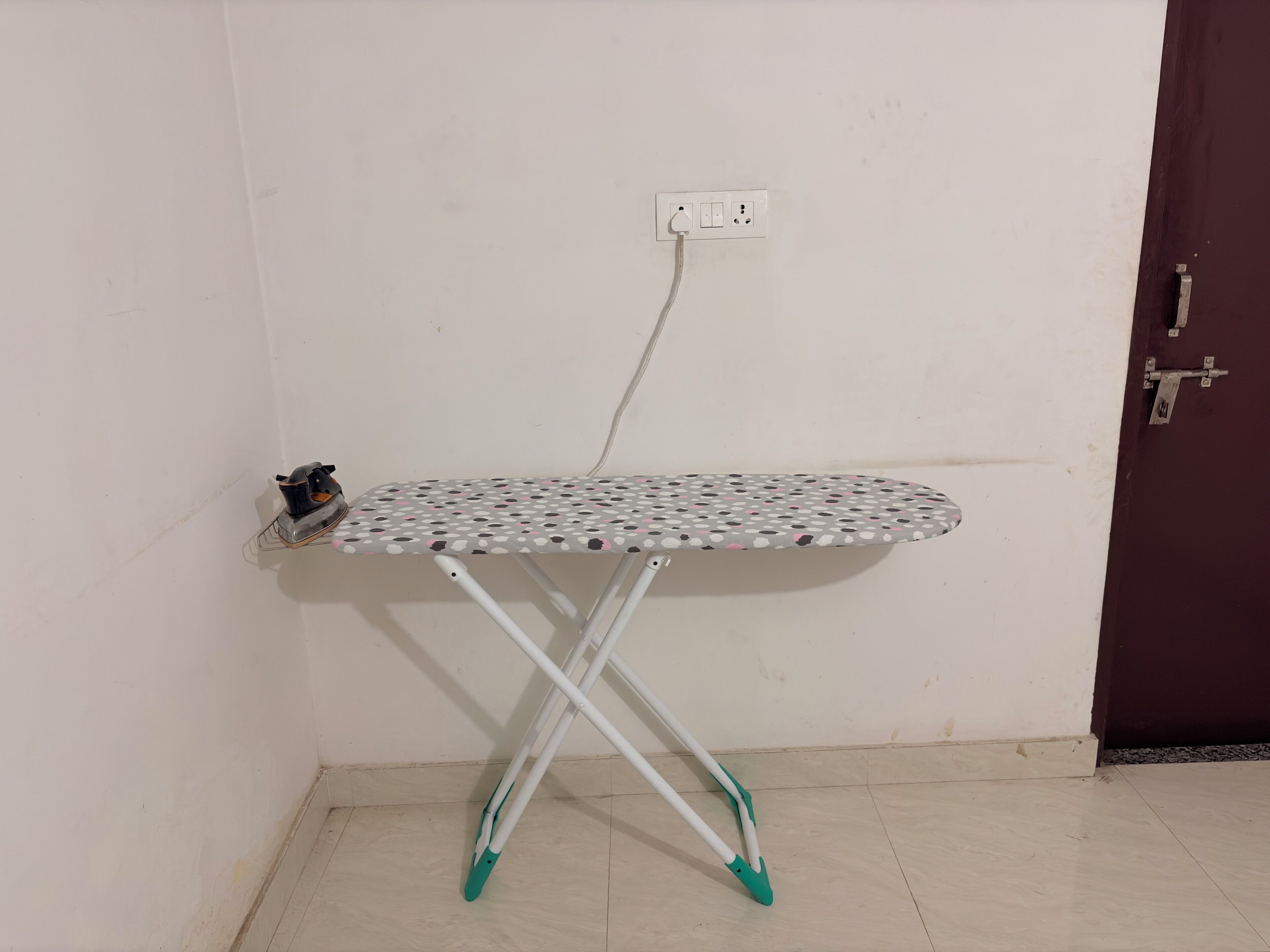 Una tabla de planchar en esquina, con un enchufe, una puerta y suelo de baldosas.