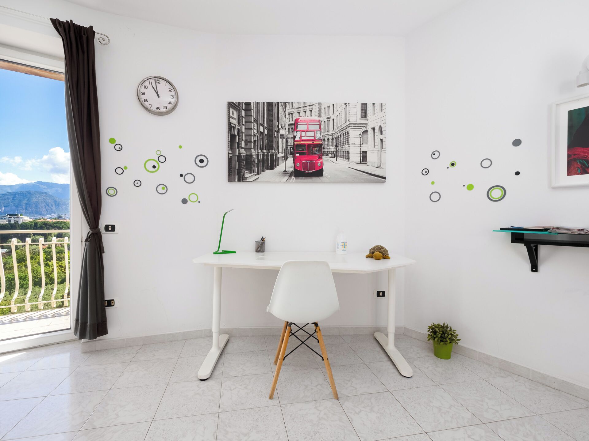 Une pièce comprenant un bureau, une chaise, une horloge, un tableau représentant un autobus rouge et donnant sur un balcon.