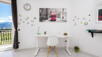 Une pièce comprenant un bureau, une chaise, une horloge, un tableau représentant un autobus rouge et donnant sur un balcon.