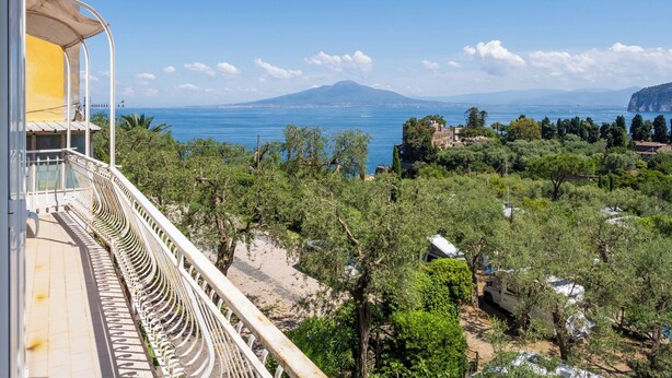 A balcony with a view of the sea, a mountain in the distance, and a building with a yellow facade. - Casa Rosa is one of those places you choose for one specific detail… and then remember for everything else. That detail is the view: a balcony opening onto the Gulf of Naples with Mount Vesuvius in front, a wide, deep panorama that accompanies every mo (Sorrento)
